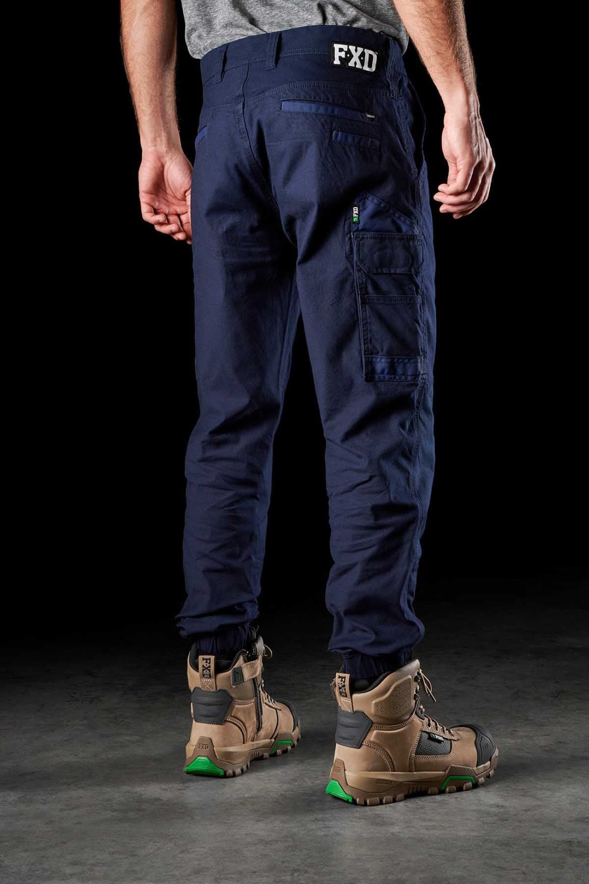 Against a dark background, a person stands with their back to the viewer, dressed in FXD WP-4 Stretch Cuffed Work Pants made of stretch cotton canvas in navy blue. The pants feature multiple pockets and are paired with brown work boots that have green soles. The waistband of the pants displays the letters "FXD" on the back.