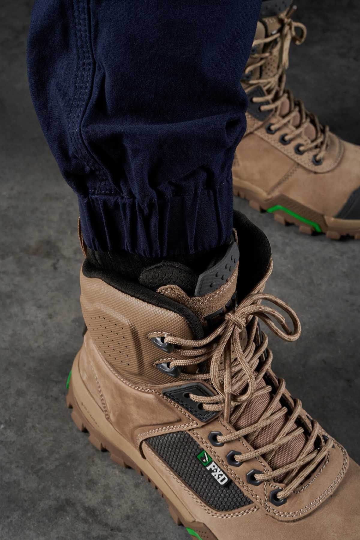 A close-up view of a person wearing tan hiking boots with thick soles and green accents. The individual is dressed in dark blue FXD WP-4 Stretch Cuffed Work Pants, with the trousers tucked into the boots made from stretch cotton canvas. The image is taken from ground level on a gray concrete surface.