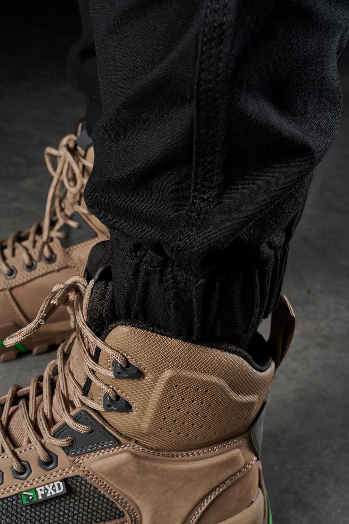 Close-up of a person wearing tan and black hiking boots with green accents, paired with FXD Womens WP-4W black cotton twill stretch work pants. The pants are tucked into the boots, showcasing the laced-up detail and sturdy design of the footwear. The image suggests readiness for outdoor activity or hiking.