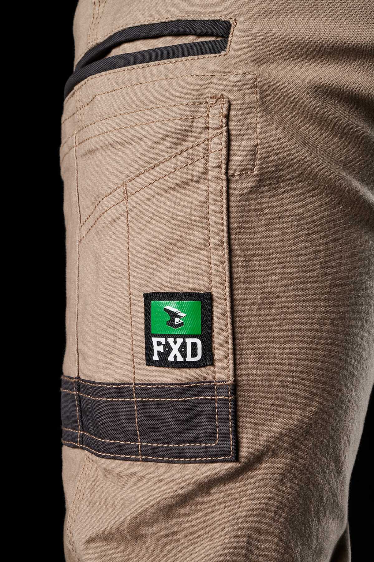 Close-up shot of the FXD Womens WP-4W beige stretch work pant pocket with black trim, featuring a green and black FXD brand patch with a white logo. Made from durable cotton twill, the pants have visible stitching and a pocket zipper. The background is black.