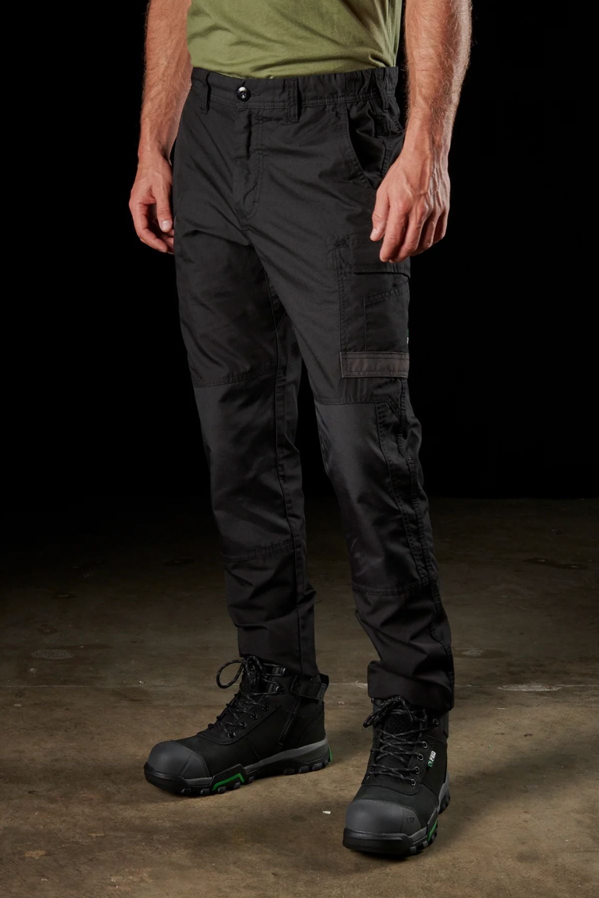 Full-body view of a person wearing the FXD WP-5 black cargo pants made from advanced stretch fabric and black work boots with green accents, standing against a plain dark background. The person is also wearing a green shirt, though only the lower part is visible. The setting appears to be indoors.