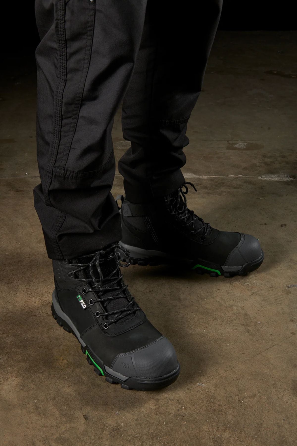 A person wearing black tactical pants featuring modern utility pockets stands on a concrete surface, complemented by the FXD WP-5 rugged work boots, which are lace-up with reinforced toes and thick soles designed for heavy-duty use. The boots feature distinct green accents.