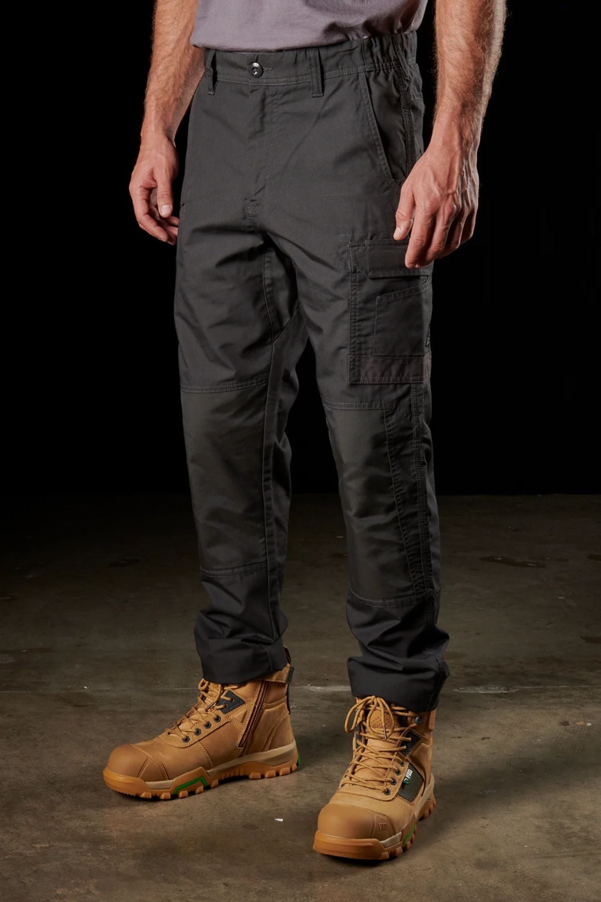 A person standing on a concrete floor, wearing FXD WP-5 dark green cargo pants made of advanced stretch fabric and tan work boots with green laces. The person's hands are at their sides, and the focused lighting highlights the texture and details of the ultra-durable FXD WP-5 pants and boots.