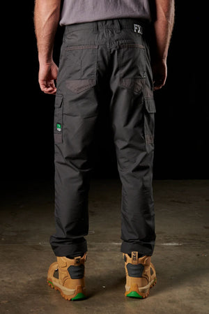 The scene features a man viewed from behind, dressed in FXD WP-5 dark cargo pants made of ultra-durable, advanced stretch fabric and a gray shirt. His legs are straight as he stands on a concrete surface, wearing rugged tan work boots with green soles against a dark background.