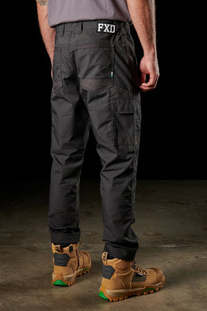 A person wearing FXD WP-5 black cargo pants, crafted from ultra-durable, advanced stretch fabric with "FXD" emblazoned on the back waistband and sturdy brown work boots, stands on a concrete floor. The individual is facing away, highlighting the rear and side pockets of the pants against a dark background.