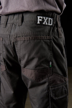 A close-up view of a person wearing the ultra-durable FXD WP-5 work pants, crafted from advanced stretch fabric for ultimate breathability, with multiple pockets and a visible "FXD" logo on the waistband. The person is standing with their back facing the camera.