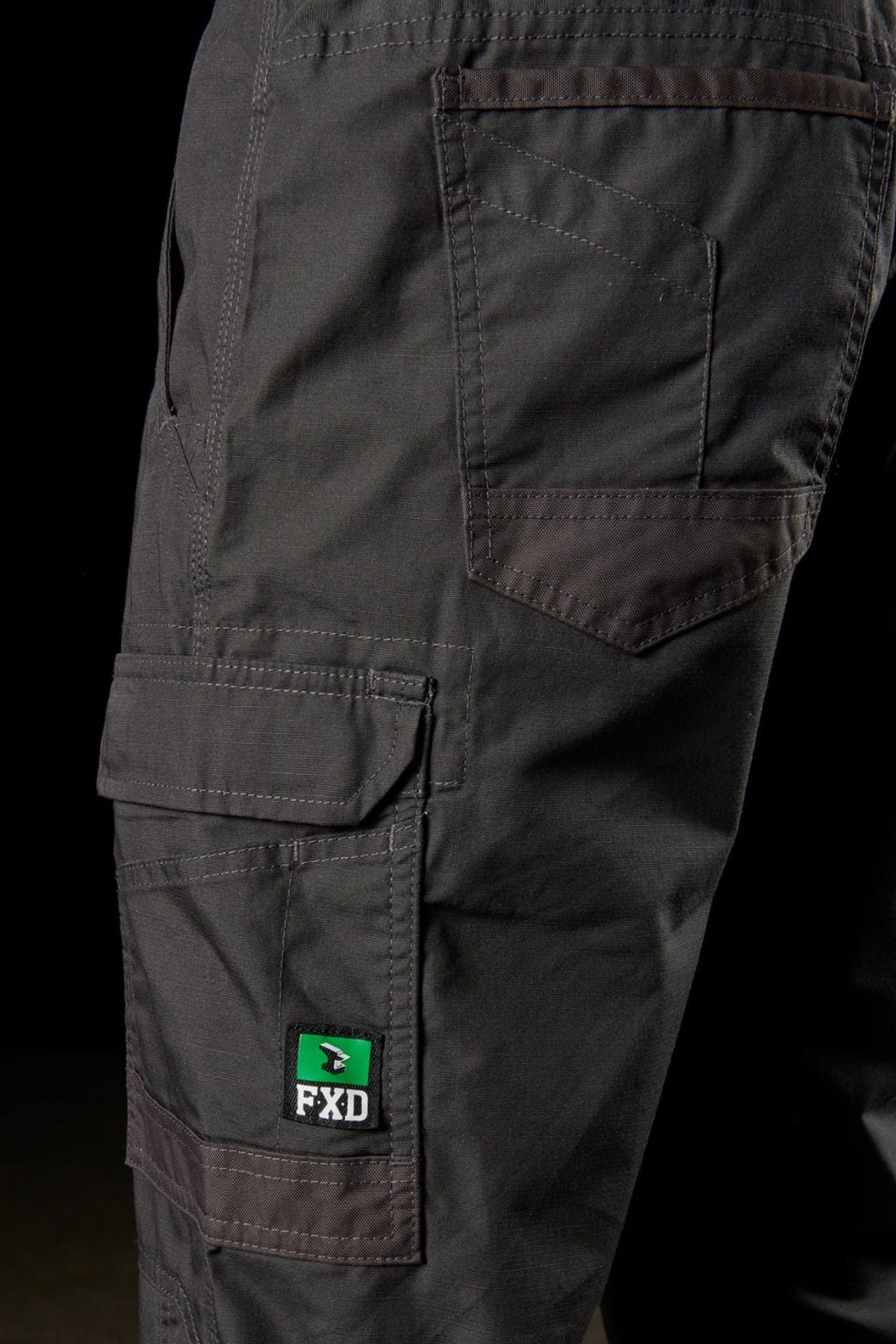 Close-up image of the FXD WP-5 dark-gray work pants featuring multiple pockets made from ultra-durable advanced stretch fabric. A green and white label with the text "FXD" is visible on the side pocket. The stitching details and texture of the fabric are highlighted against a black background.