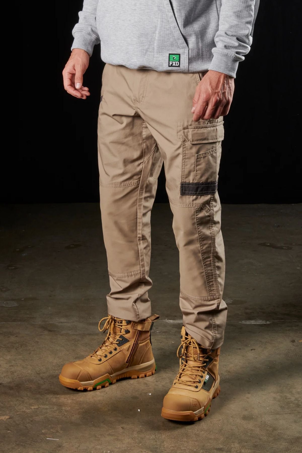 The lower half of a person is shown standing against a dark background, wearing the FXD WP-5 khaki cargo pants with modern utility pockets, paired with brown work boots featuring green accents. A light grey hoodie and an ID badge clipped to the sweatshirt complete the outfit.