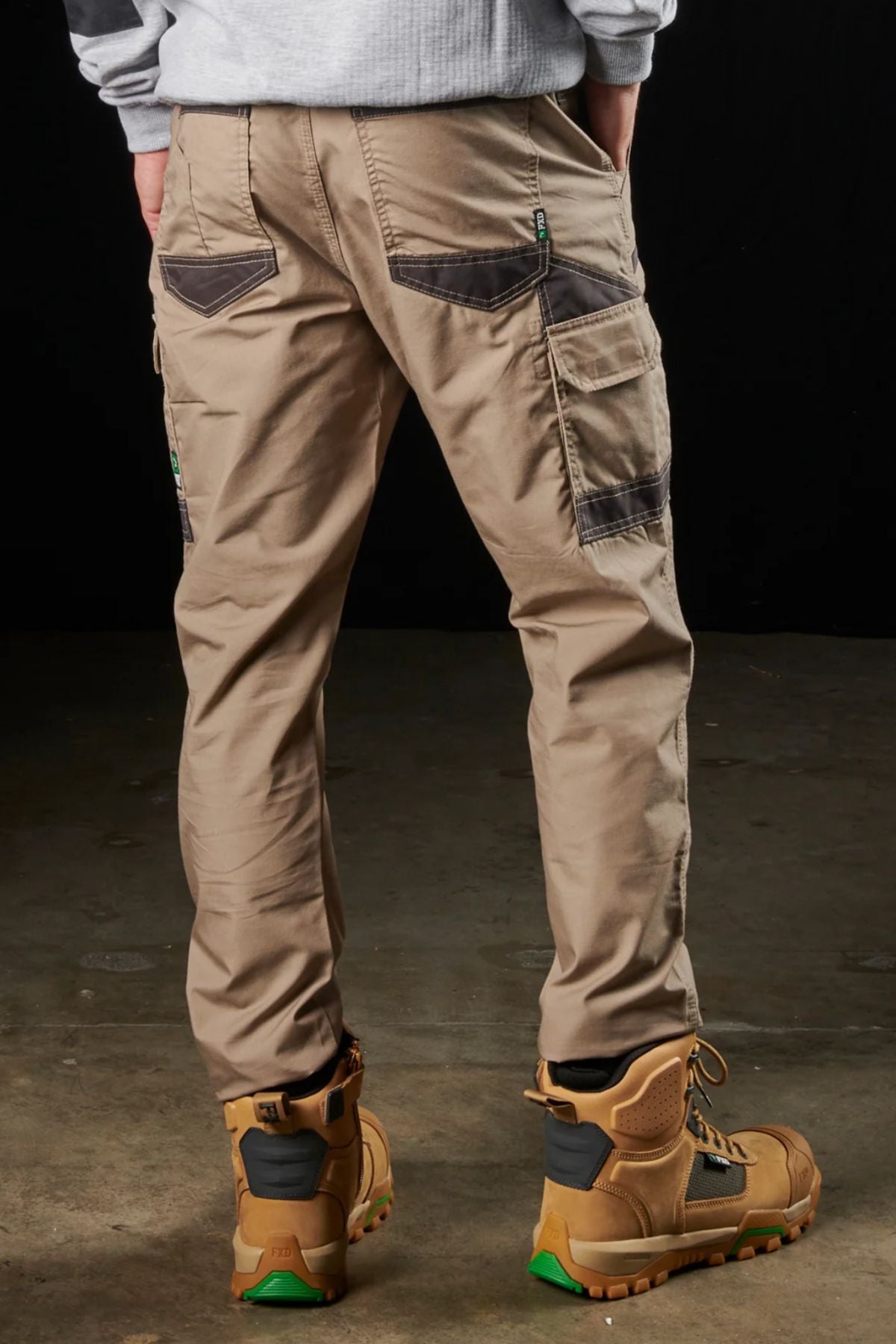 A person stands with their back facing the camera, dressed in tan FXD WP-5 work pants crafted from advanced stretch fabric and equipped with modern utility pockets, complemented by brown-and-black work boots. The backdrop is dark, and the floor seems to be a concrete or industrial surface.
