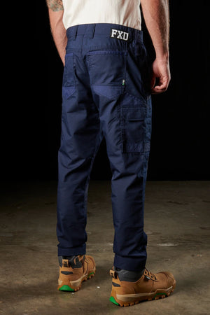 A person stands on a concrete floor, facing away from the camera. They are clad in a white shirt, navy blue FXD WP-5 cargo pants featuring multiple pockets and brown work boots with green accents. The FXD WP-5 pants provide ultimate breathability and advanced stretch fabric for maximum comfort.