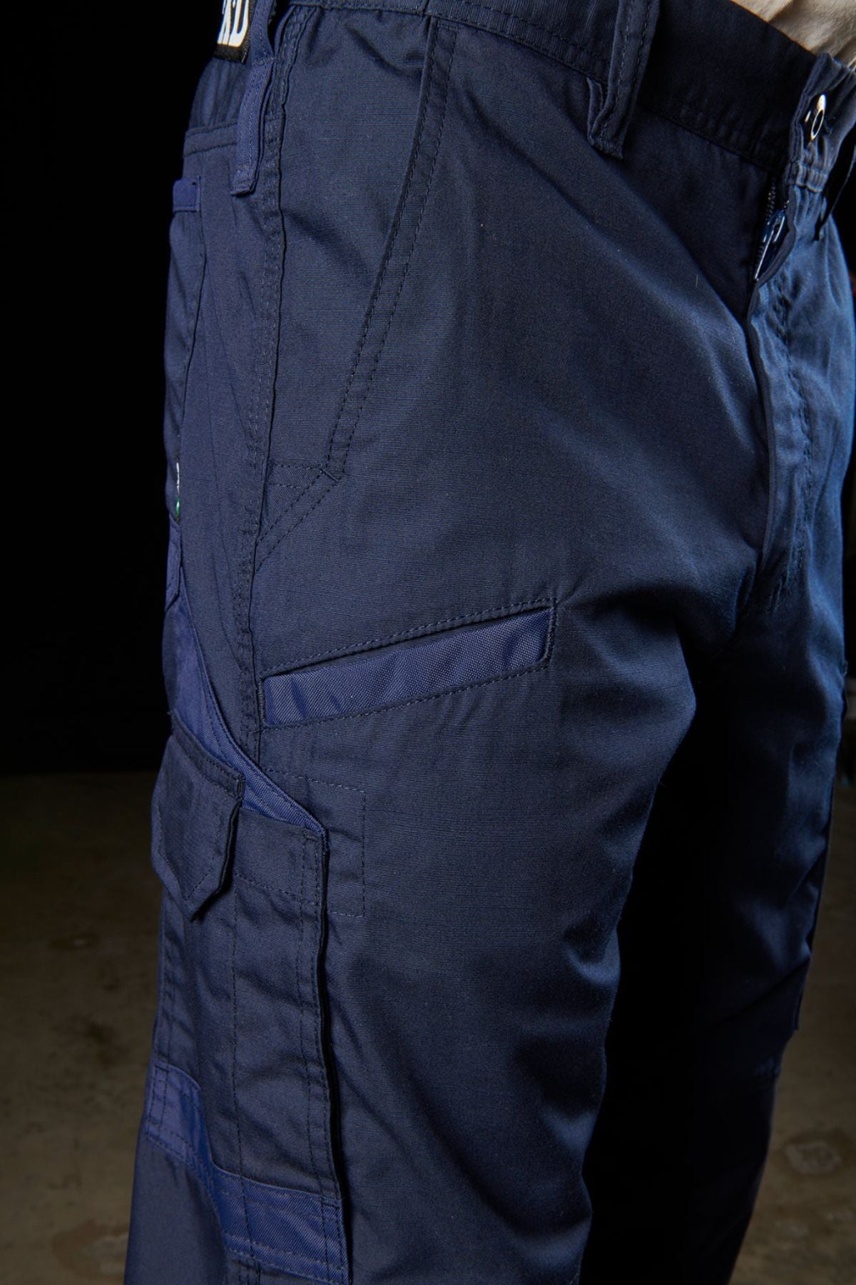 A close-up of a person wearing FXD WP-5 cargo pants in navy blue showcases the multiple pockets on the left side, including a flap pocket and a zippered pocket. The durable advanced stretch fabric is clearly visible, set against a dark background that makes the pants stand out prominently.