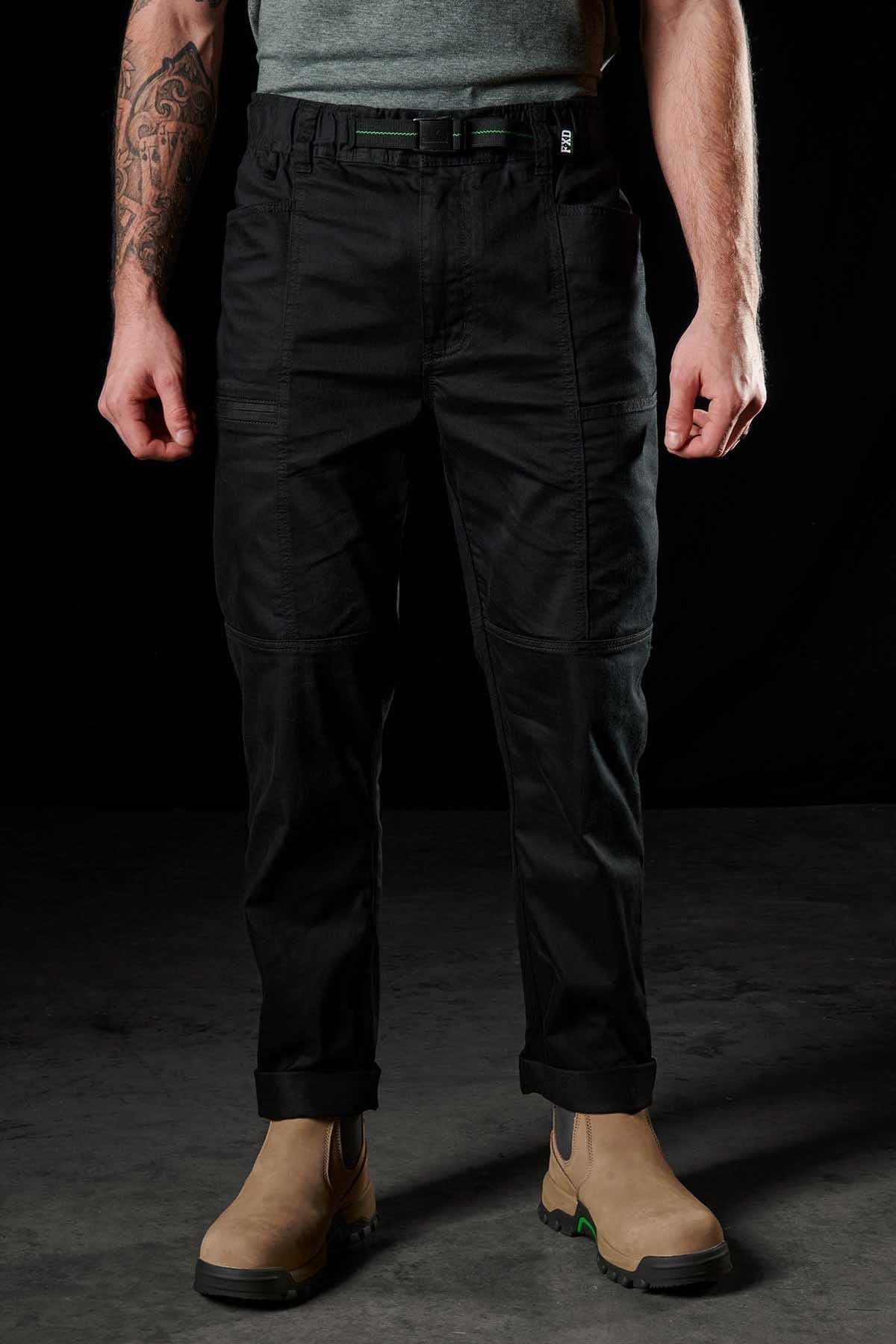 A person in FXD WP-6 black work pants with a stretch waistband, a grey t-shirt, and tan utility boots stands against a dark backdrop, revealing tattoos on one arm.