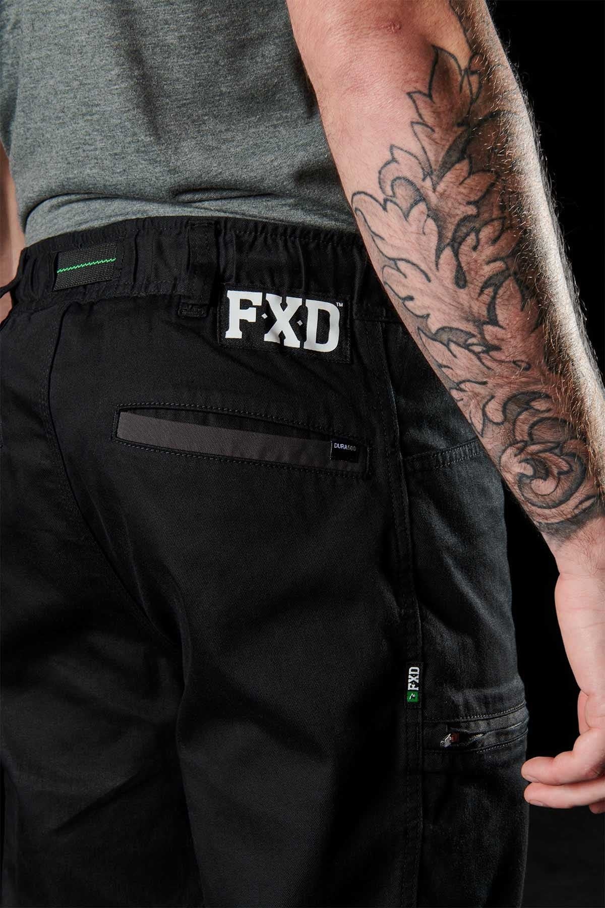 A person with a tattooed arm is wearing black FXD WP-6 work pants featuring a stretch waistband and visible pockets, plus an "FXD" label on the back. The partially visible gray shirt completes an outfit designed for comfort and durability.
