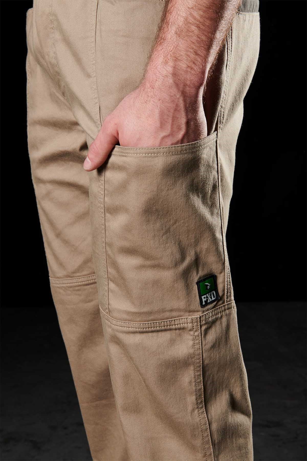 A close-up of a person’s arm resting in the pocket of FXD WP-6 tan work pants with stretch waistband, reinforced seams, and a green FXD logo patch on the thigh, set against a black background.