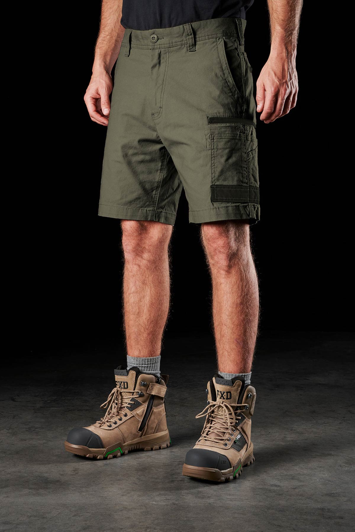 A man wearing FXD WS-3 Short stretch work shorts, a black shirt, gray socks, and tan boots stands on a dark surface against a black background. Only his legs and lower torso are visible.
