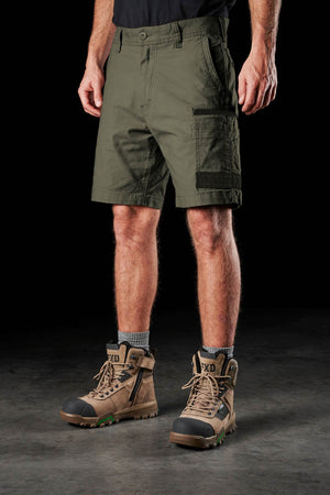 A man wearing FXD WS-3 Short stretch work shorts, a black shirt, gray socks, and tan boots stands on a dark surface against a black background. Only his legs and lower torso are visible.