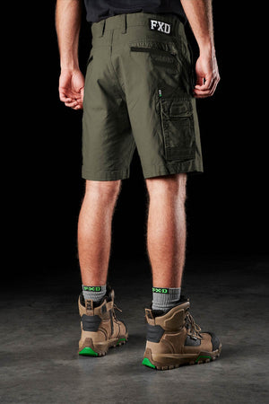 Seen from behind on a dark floor, a person wears olive green FXD WS-3 Shorts made of stretch cotton canvas, paired with gray FXD socks and brown work boots featuring green accents.