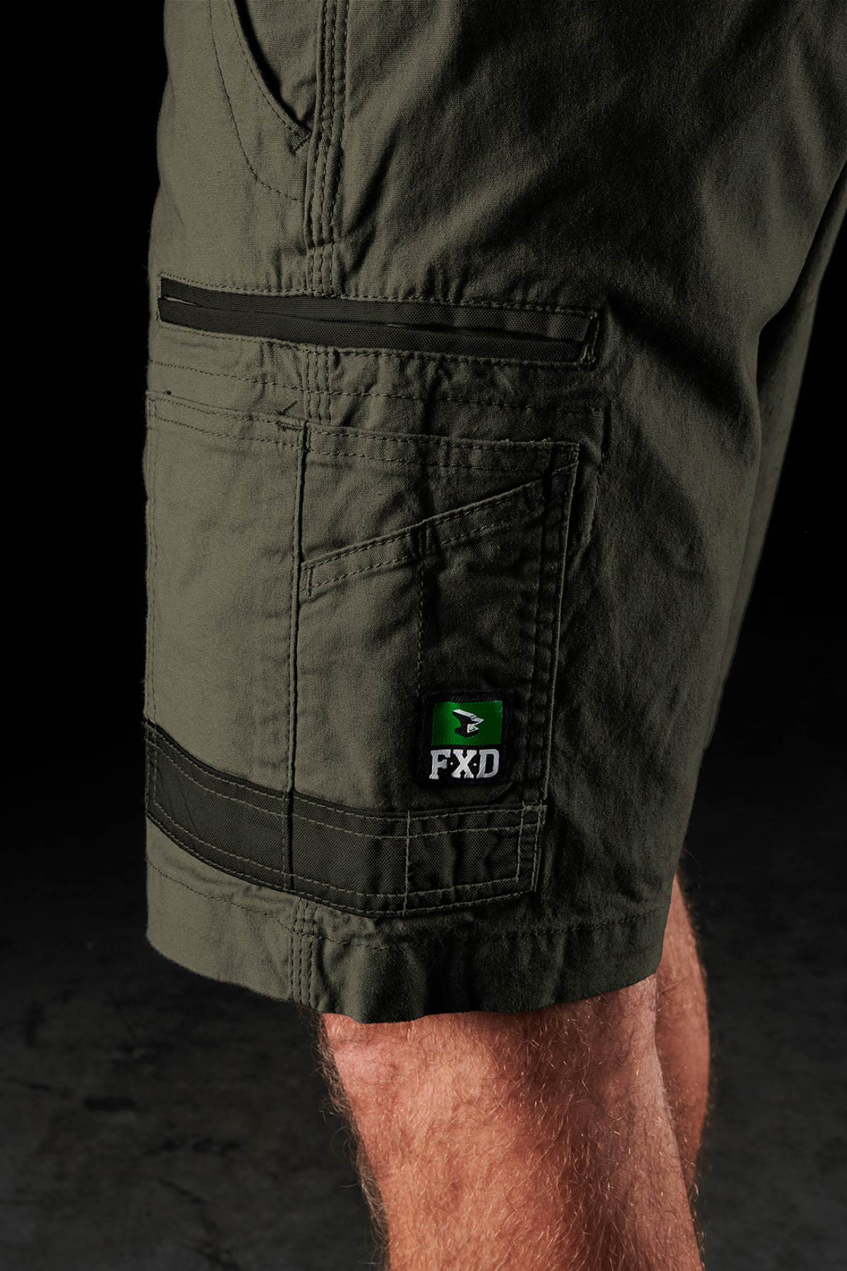 A close-up of a person wearing FXD WS-3 Short—durable stretch cotton canvas work shorts with contrast stitching, a side pocket, and an "FXD" logo patch above the knee. The background is dark and out of focus.