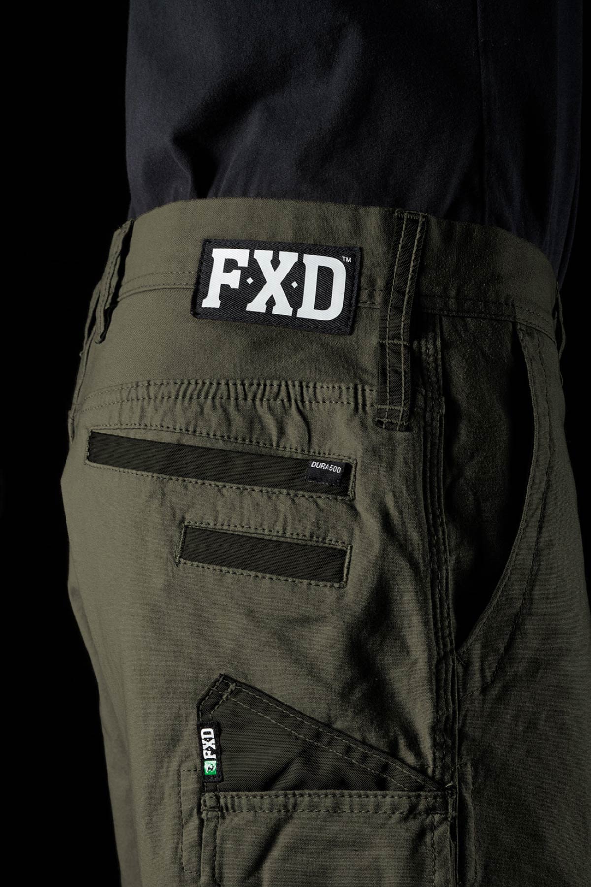 Close-up of a person in olive green FXD WS-3 Short stretch cotton canvas shorts and a black shirt, with the FXD logo patch above the back pocket and detailed stitching highlighting the durable design.