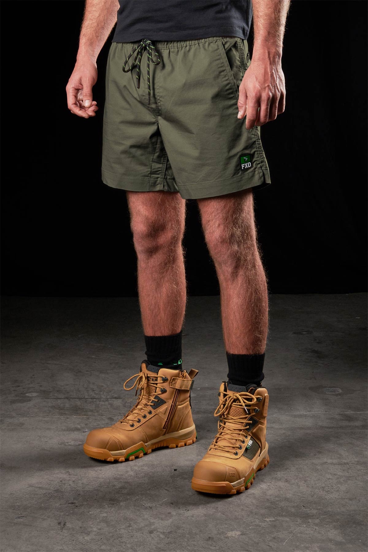 A person wearing FXD WS-4 olive green stretch ripstop work shorts, a black T-shirt, black socks, and tan lace-up boots stands on a concrete floor against a black background.