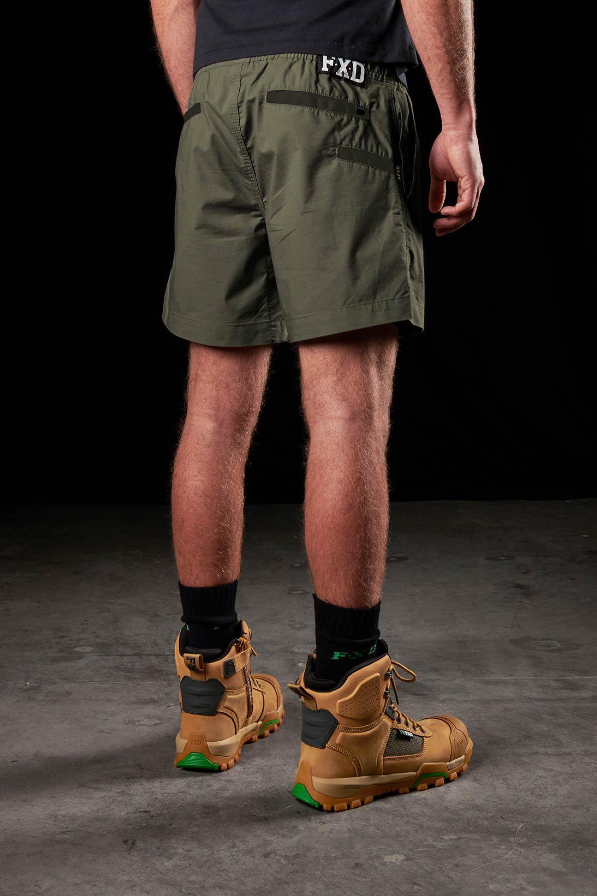 A person stands on a concrete floor, facing away from the camera, wearing olive green FXD WS-4 Shorts, black socks, and tan work boots with green accents against a black background.
