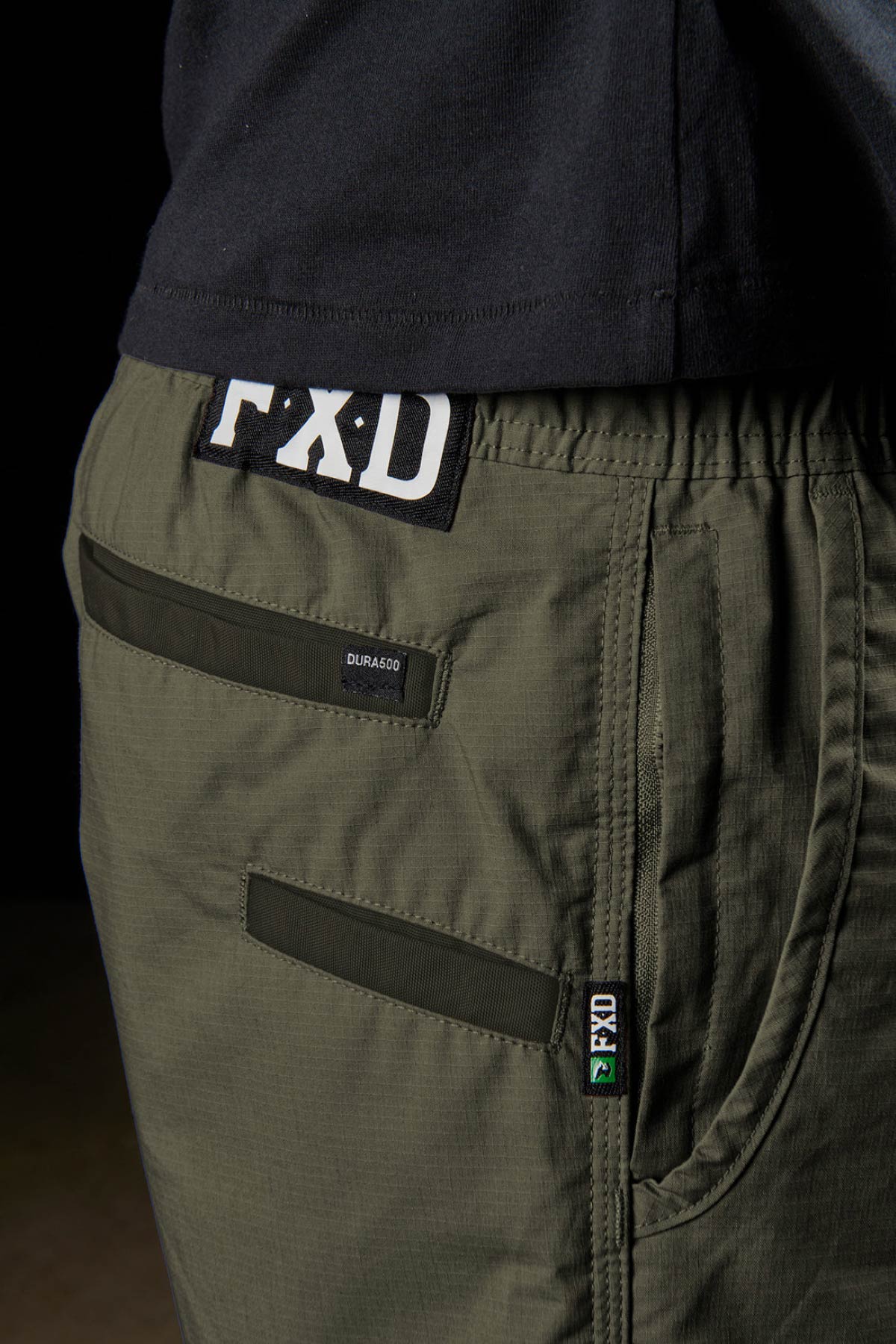 Close-up of a person wearing olive green FXD WS-4 Shorts with elastic waistband, black label tags, and a black shirt partially visible. Focus is on the back and side pocket details of the FXD shorts.