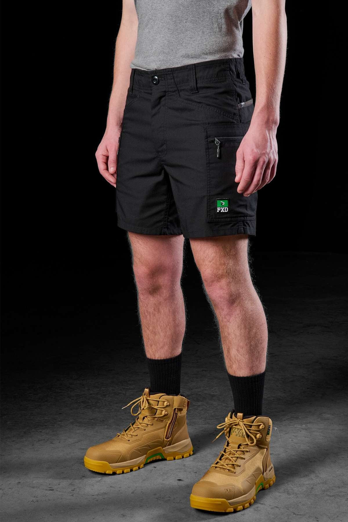A person dressed in a gray shirt, black FXD WS-6 Work Short emblazoned with a patch, black socks, and tan work boots demonstrates the stretch mobility feature. The dark backdrop accentuates the practical style of the outfit.