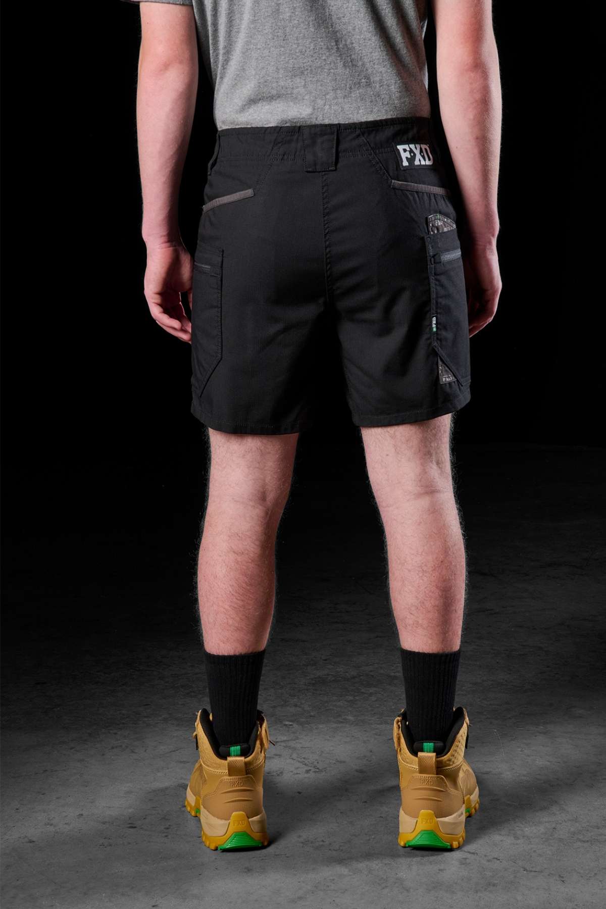 A person stands on a concrete floor wearing a grey t-shirt and the FXD WS-6 Work Short, known for its stretch mobility. Black socks and tan work boots complete the ensemble. The black background highlights the rear view of this functional outfit, designed for both work efficiency and style.