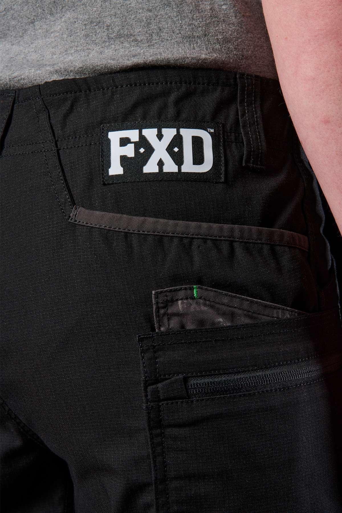 A close-up shot of the black FXD WS-6 Work Short's back pocket area highlights a prominent FXD logo. The shorts are designed with reinforced stitching, stretch mobility, and include a convenient side pocket with a zipper. The durable texture suggests they're made for demanding work environments.