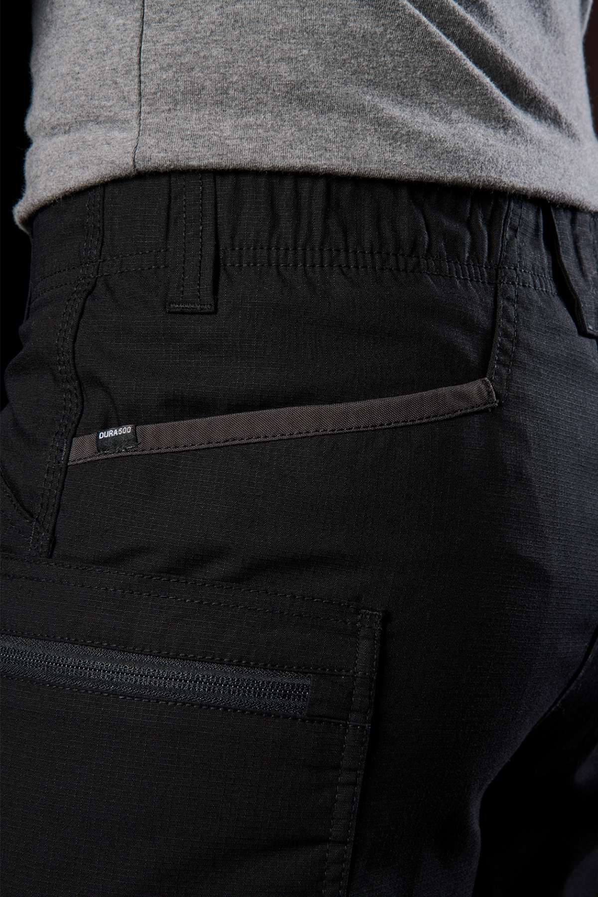 A close-up shows someone wearing black pants with zippered pockets, crafted for stretch mobility and durability. The fabric texture pairs well with a grey shirt, echoing the practical style of FXD WS-6 Work Shorts or traditional cargo shorts.