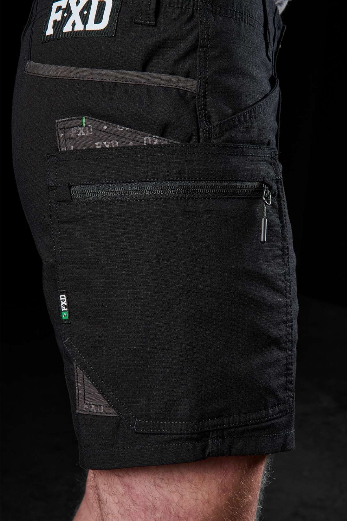A detailed view features an individual sporting the FXD WS-6 Work Short, highlighting its black cargo design equipped with multiple pockets, a zipper, and "FXD" branding. The shorts are noted for their stretch mobility, intricate stitching, and rugged texture set against a dark backdrop.