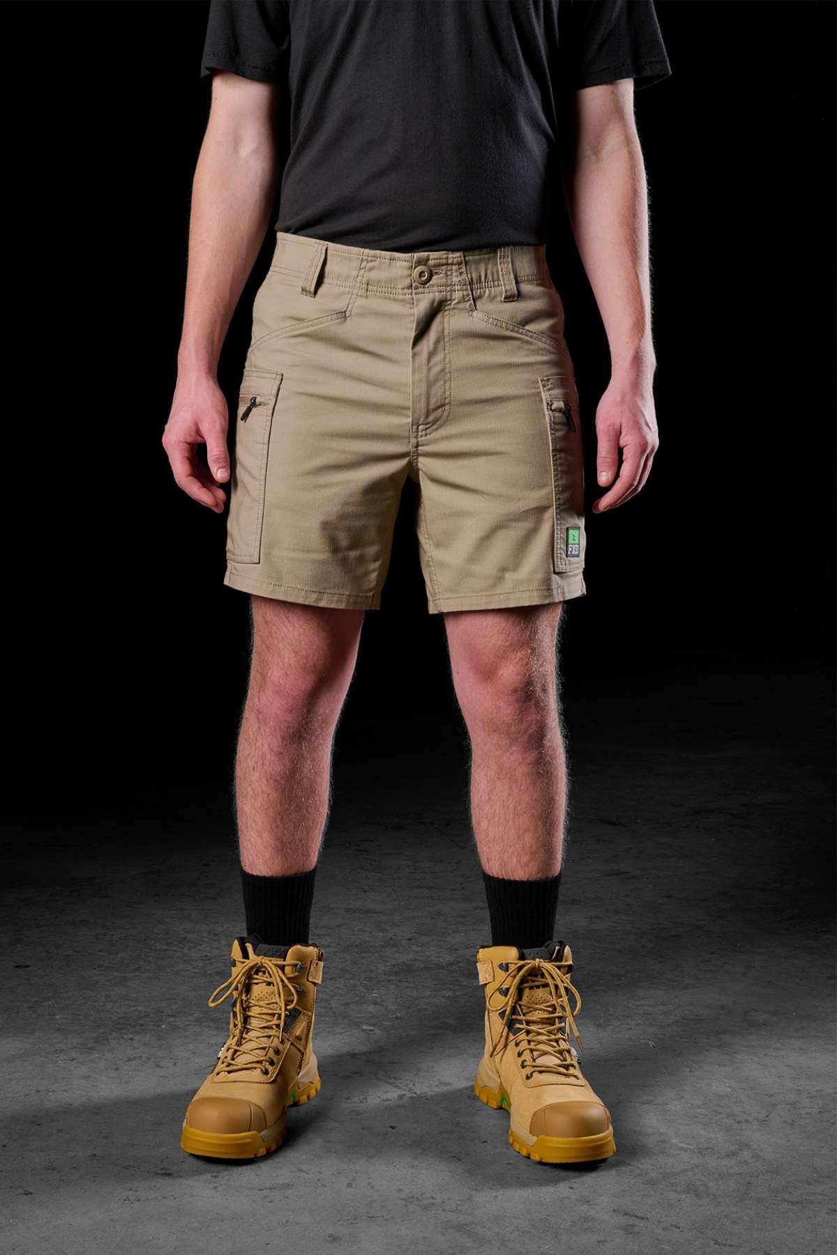 A person stands against a dark background, showcasing comfort and practicality in a black t-shirt, FXD WS-6 Work Short in khaki, and tan work boots. The shorts feature spacious pockets, with their hands resting casually at their sides.