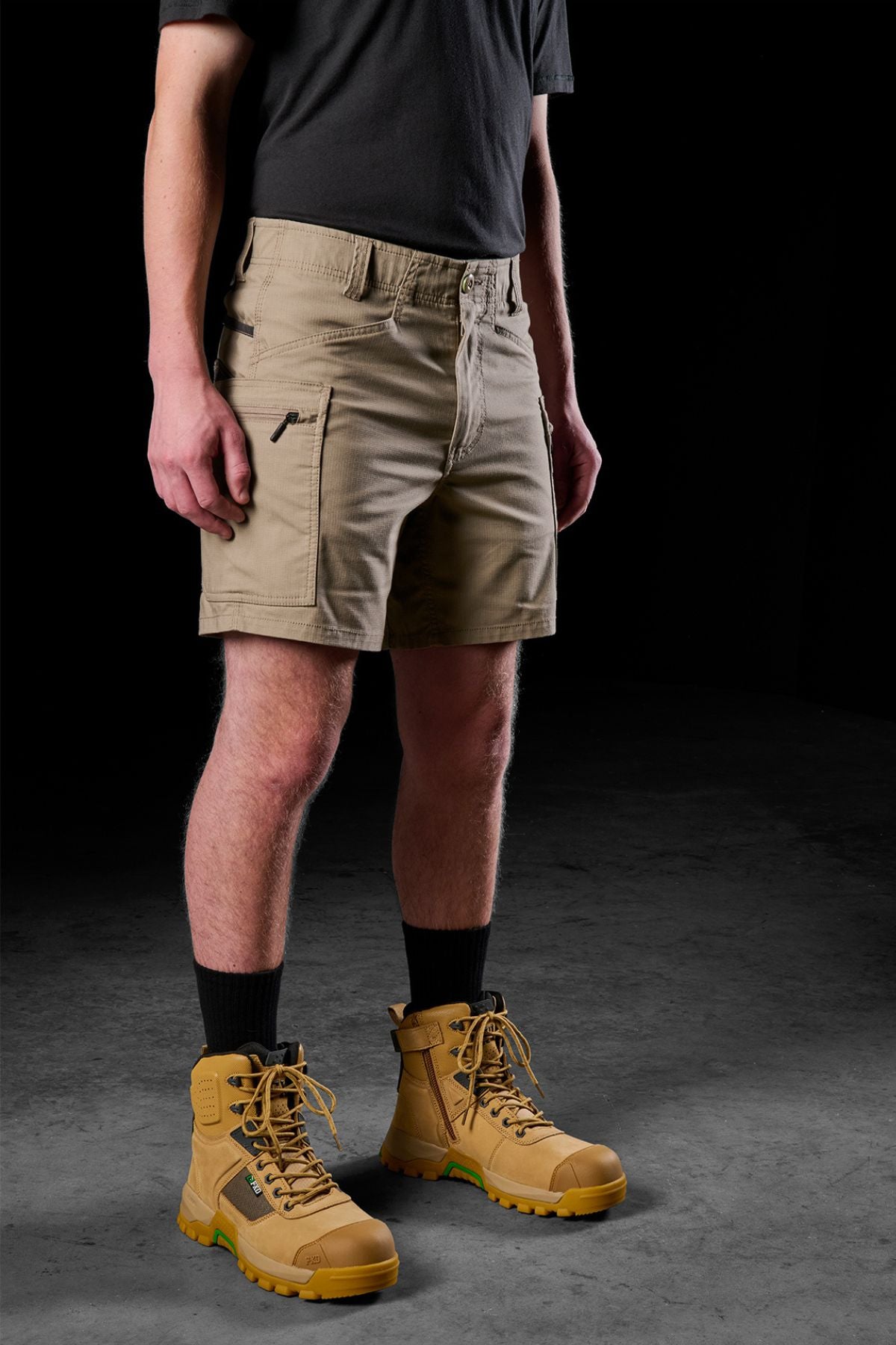 A person dressed in a black shirt and FXD WS-6 Work Shorts stands confidently in yellow lace-up boots, complemented by thick black socks. The dark background highlights the comfort and practicality of their casual attire and durable footwear.