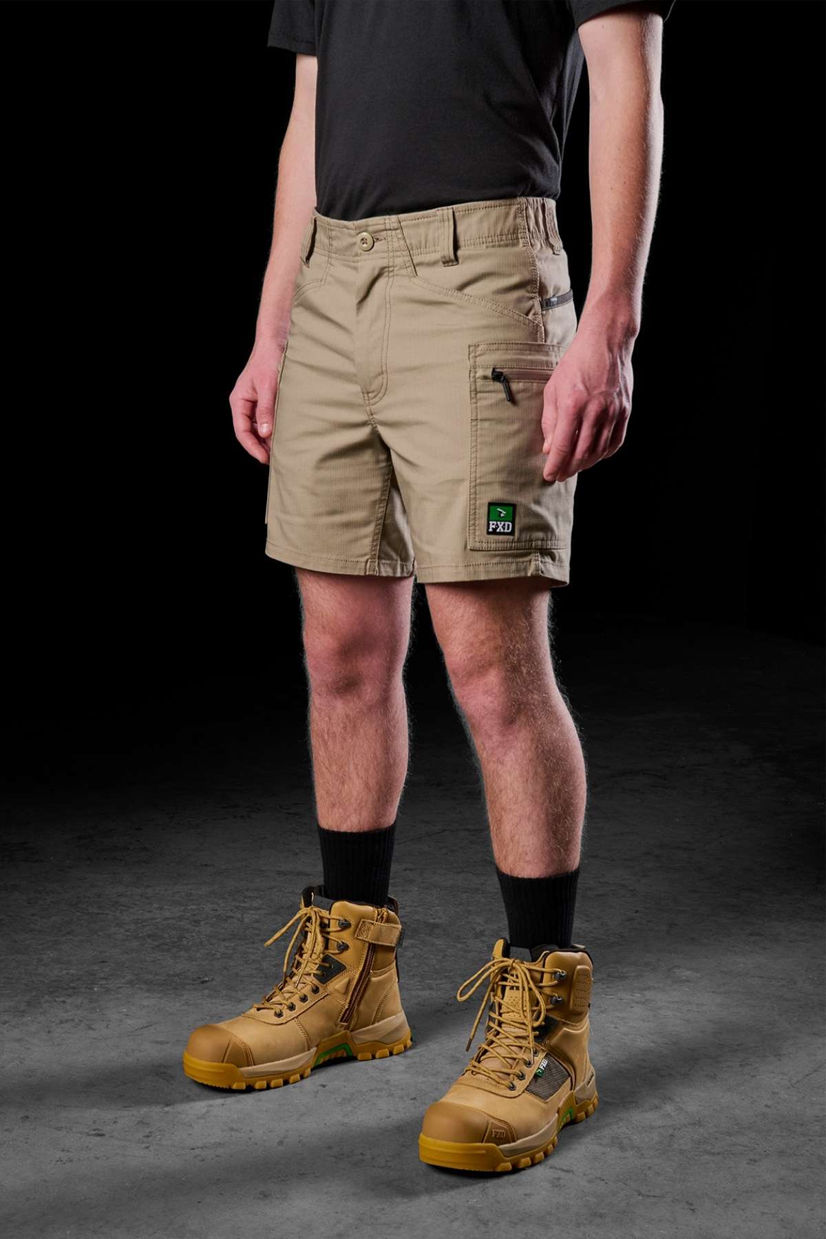 Dressed in a black t-shirt, the FXD WS-6 Work Short adorned with a green "Pro" patch, black socks, and tan work boots, an individual stands against a dark background, seamlessly merging comfort with practicality.
