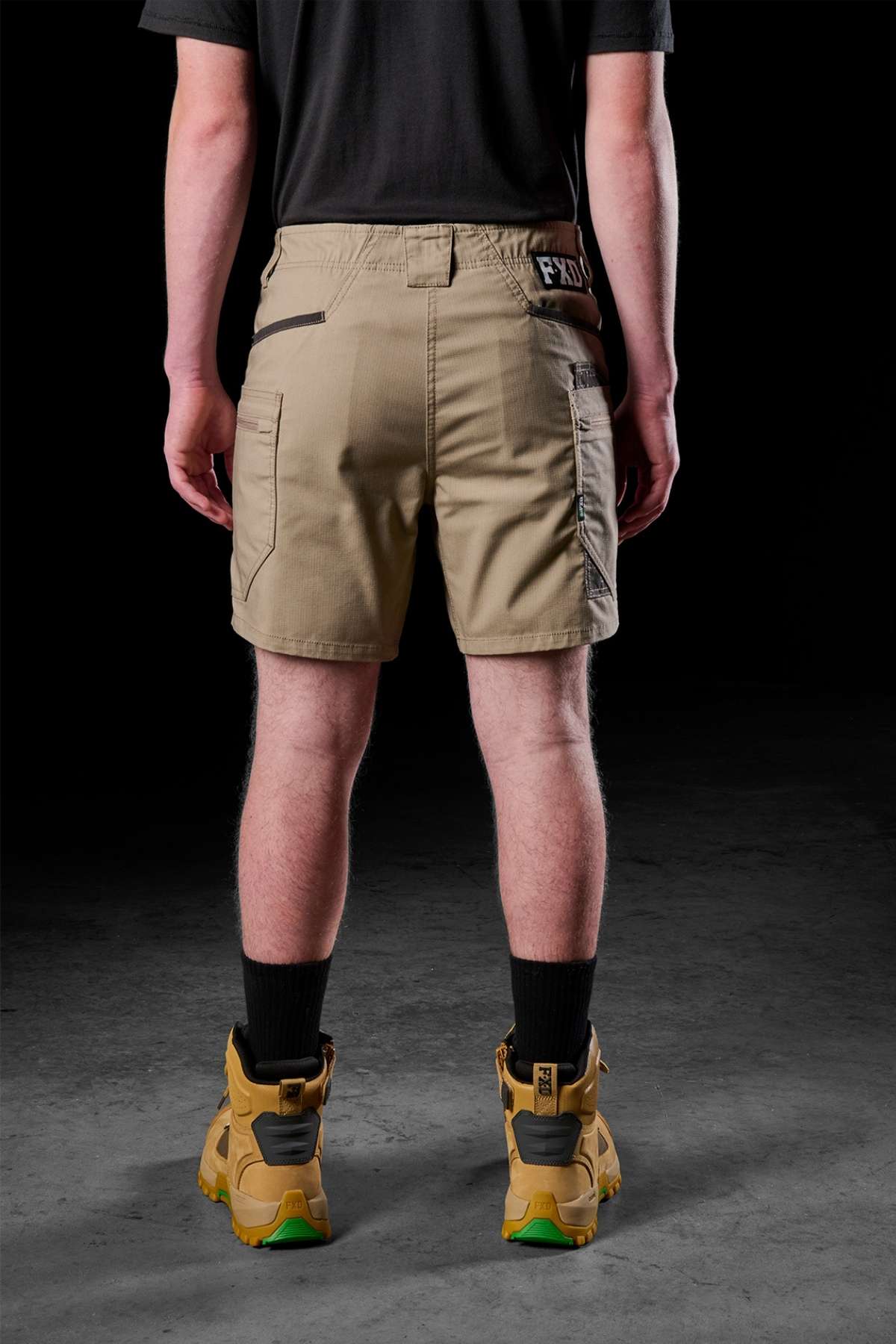 A person dressed in a black shirt, light brown FXD WS-6 Work Shorts known for their comfort and practicality, black socks, and tan boots stands against a dark background with their back to the camera.