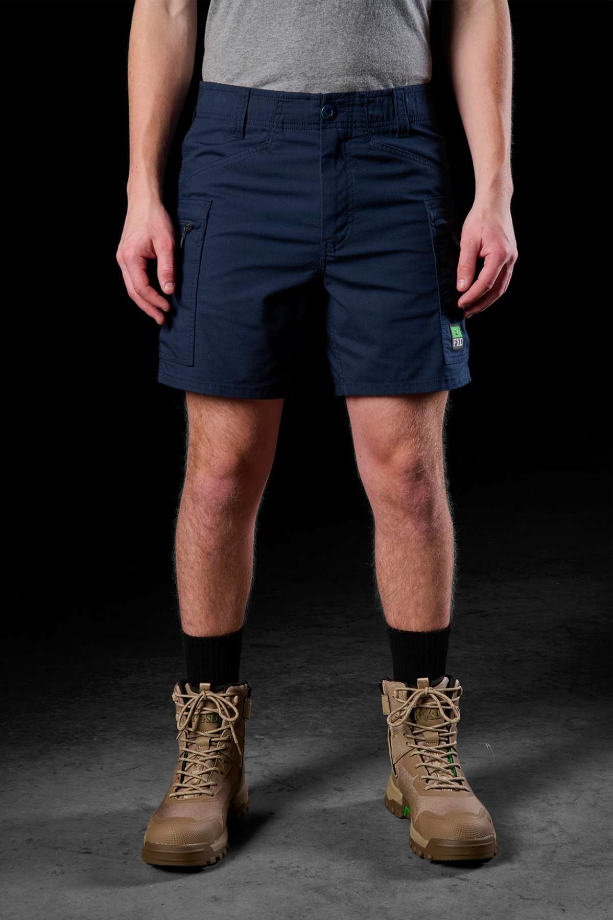 Person wearing navy FXD WS-6 Work Short with added stretch, a gray T-shirt, and brown hiking boots with black socks against a dark background.