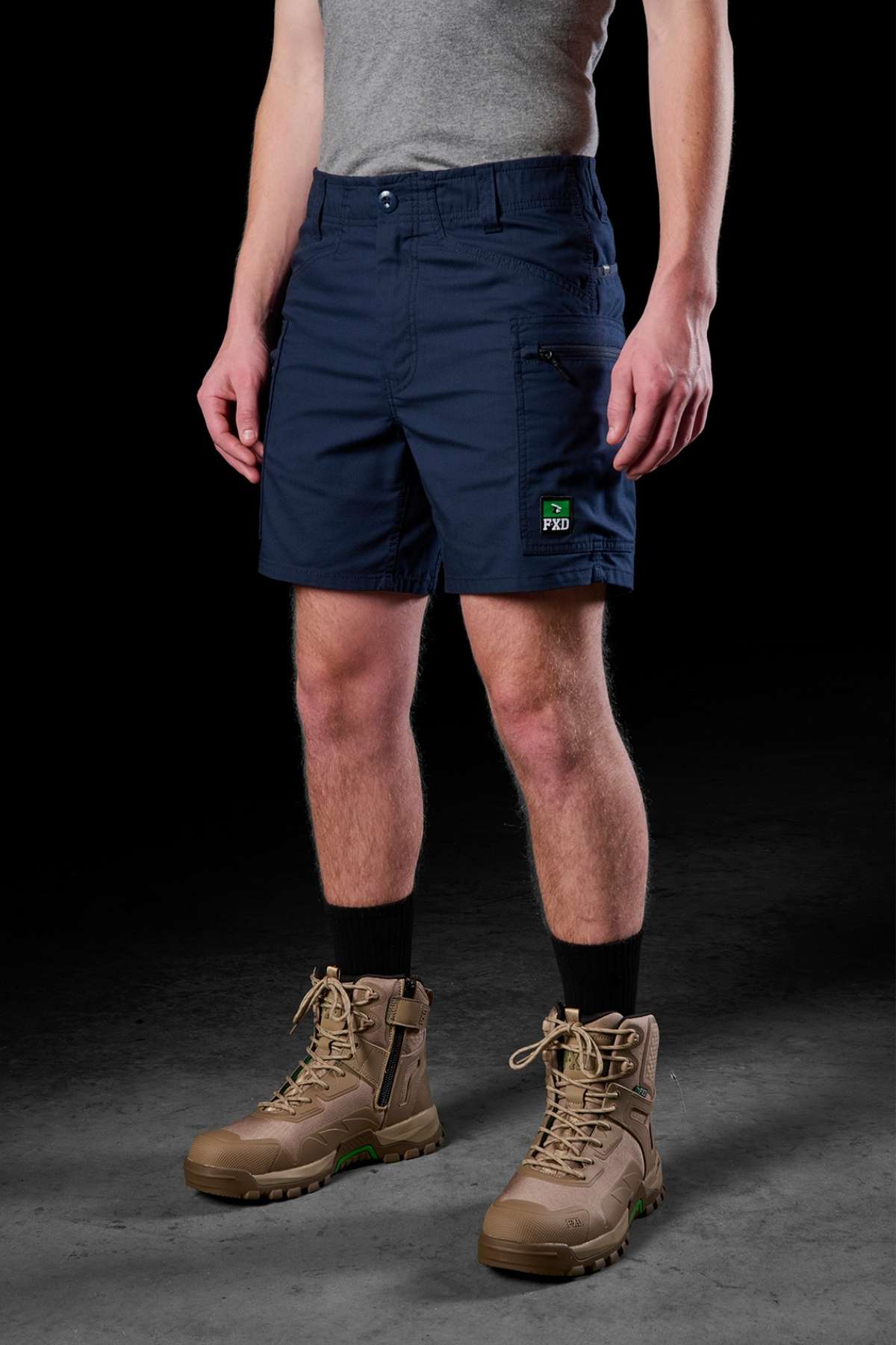 A person is wearing the FXD WS-6 Work Short in navy blue, paired with a gray T-shirt and tan tactical boots with black socks. The shorts feature a small black and green logo patch on the left side and offer added stretch for comfort. The background is dark and plain.