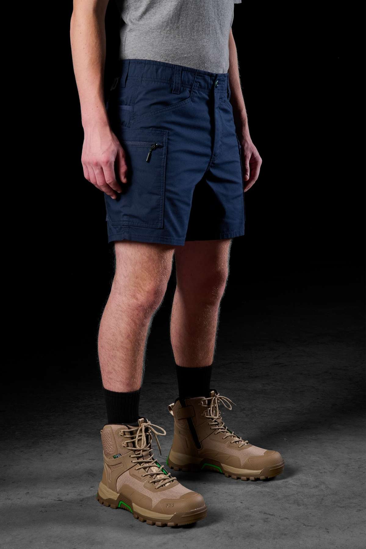 A person in a gray T-shirt and FXD WS-6 Work Shorts stands against a dark background, showcasing their outfit with blue cargo shorts paired with black socks and beige lace-up boots. This attire is designed to offer flexibility and added stretch for comfort.