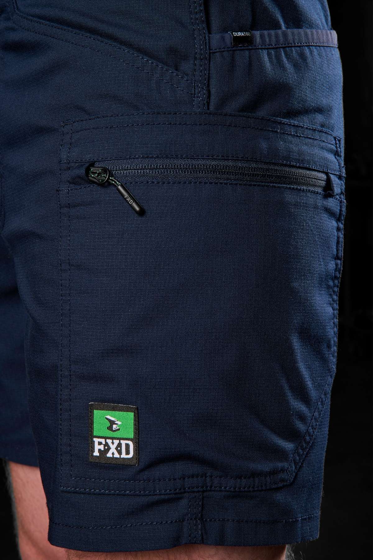 A detailed view of someone wearing the navy FXD WS-6 Work Short highlights the zippered pocket and the distinct green, black, and white "FXD" logo at the bottom. These cargo shorts boast added stretch for comfort and a textured fabric, set against a dark backdrop.