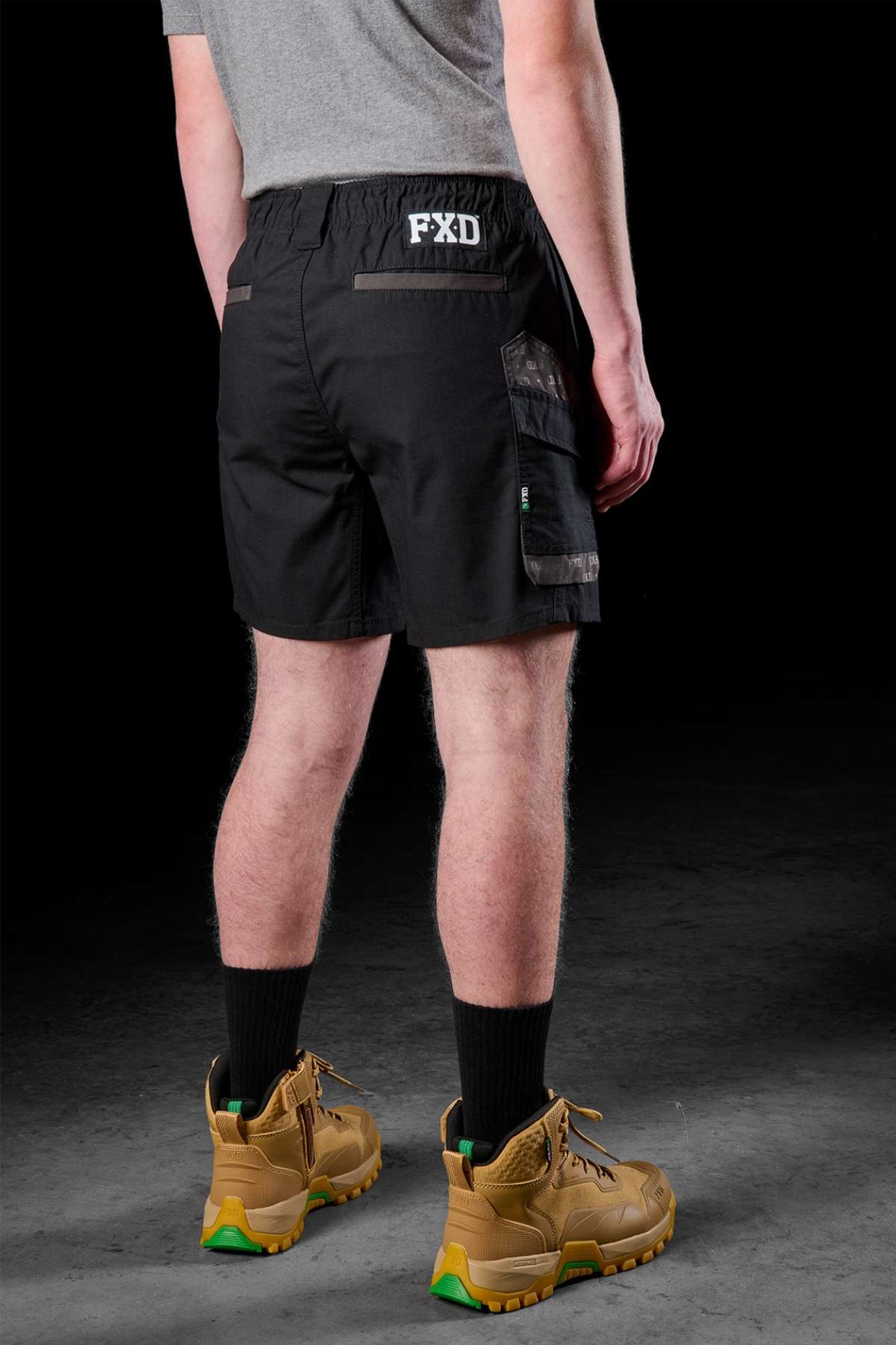 A man faces away from the camera, dressed in a grey T-shirt paired with the resilient FXD WS-7 Work Short, featuring an elastic waist. His ensemble is completed with black socks and tan work boots highlighted by green accents, all presented in front of a dark backdrop.