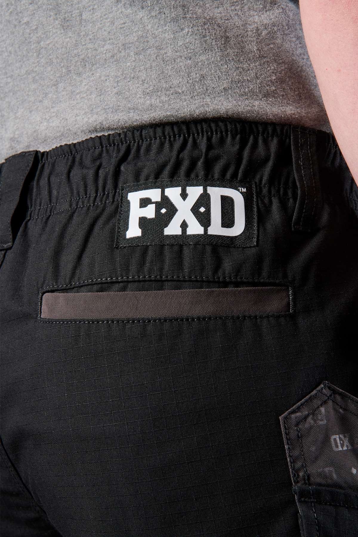 A person is wearing the FXD WS-7 Work Short in grey, designed with an elastic waistband. A black band adorns the back pocket, prominently featuring the "FXD" logo above it. The fabric looks robust, making it ideal for both work and casual wear.