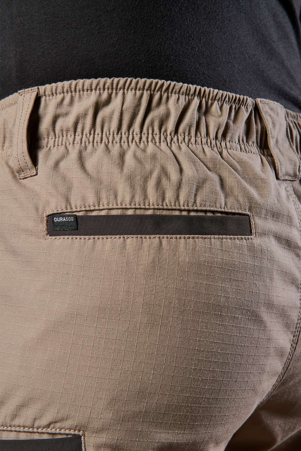 Close-up of tan cargo pants highlighting the back pocket, which features a black label and an elastic waistband. The fabric is enhanced with a subtle grid pattern and features a black loop detail beneath the pocket, providing utility and comfort similar to the FXD WS-7 Work Short.
