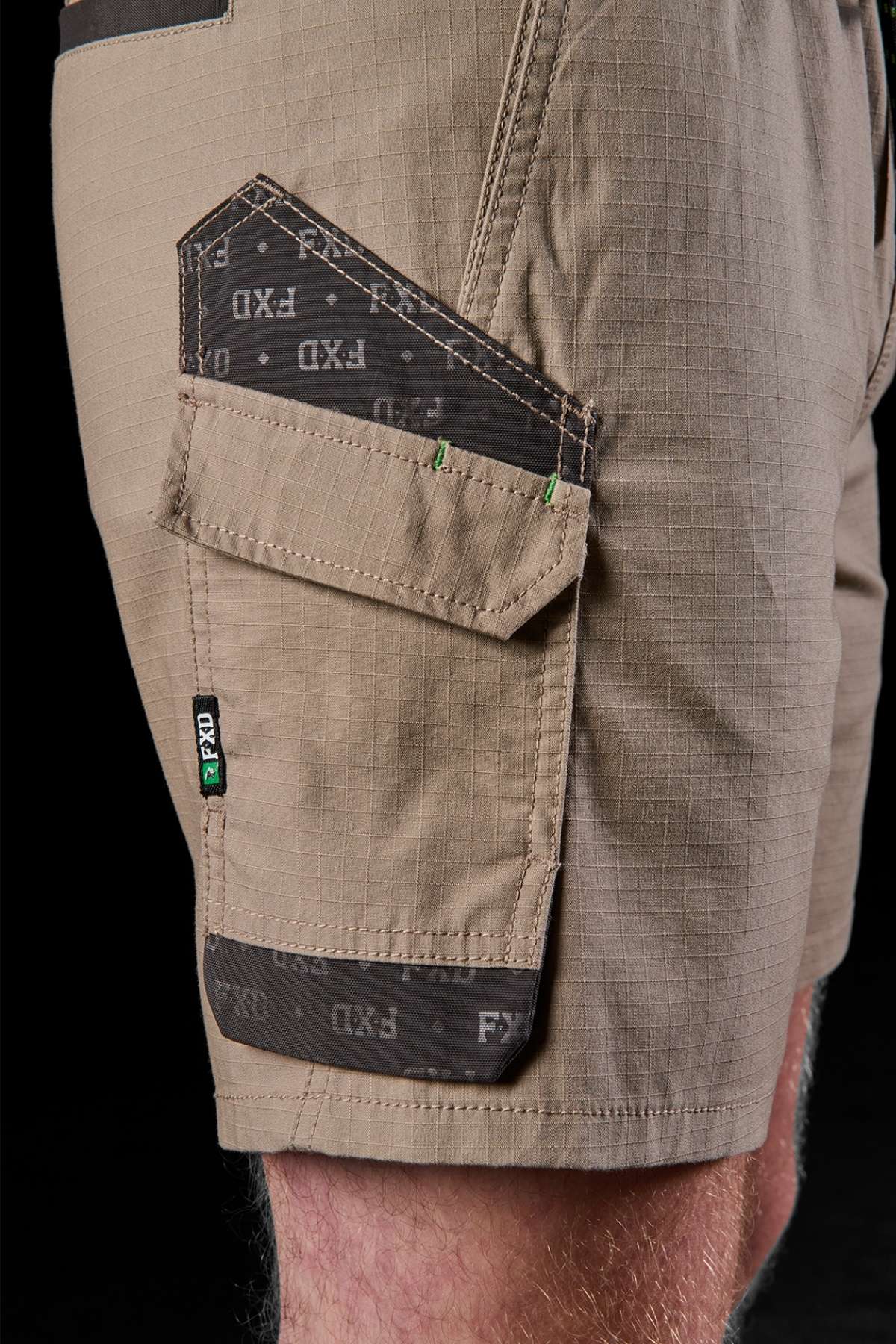A close-up shot captures a person wearing the FXD WS-7 Work Short in beige, emphasizing its comfort and practicality. The shorts showcase a black and gray pocket design adorned with the "FXD" logo, along with reinforced stitching and a flap-covered side pocket, all against a stylish black backdrop.