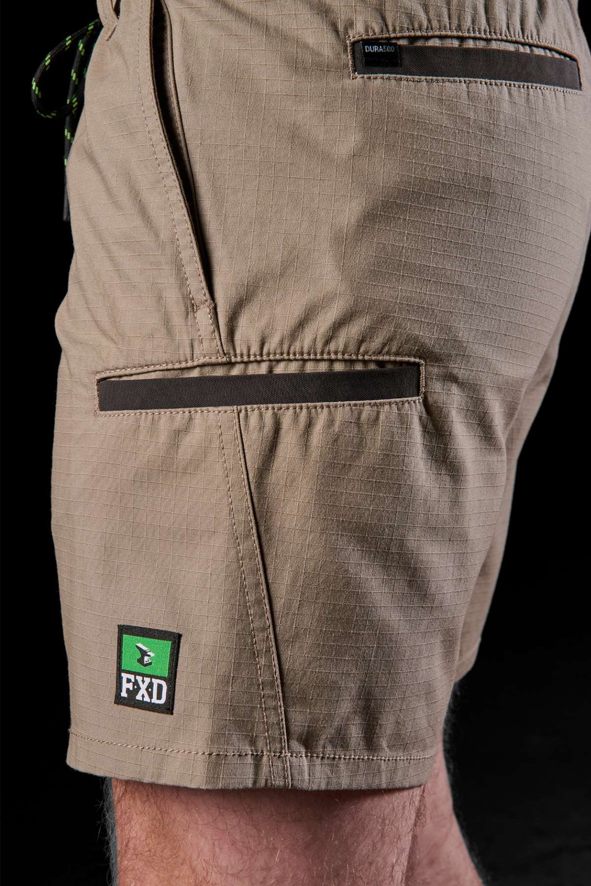 A close-up shows an individual wearing the tan FXD WS-7 Work Short, which includes zippered pockets and a green FXD logo patch on the side. The fabric is durable and slightly textured, providing comfort and functionality for daily use.