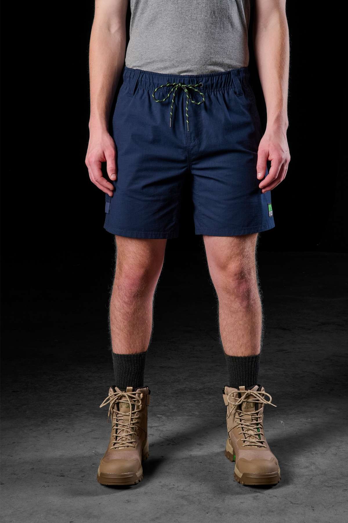 A person stands against a dark background wearing a gray t-shirt and the FXD WS-7 Work Short in durable navy blue, complete with drawstrings, complemented by tall dark socks and tan lace-up boots.