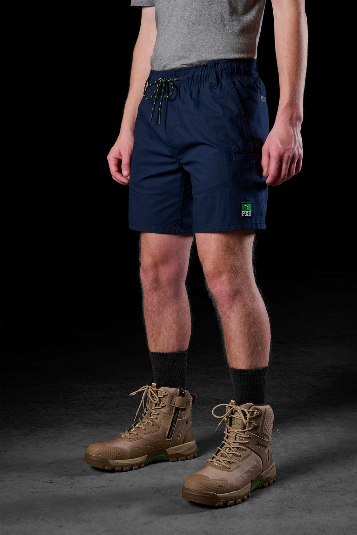 A person stands against a dark background, dressed in a gray t-shirt and FXD WS-7 Work Short, which are durable navy blue work shorts featuring a small green emblem. They complete the outfit with tan hiking boots paired with black socks. The comfortable shorts include a drawstring, and the individual's hands rest by their sides.
