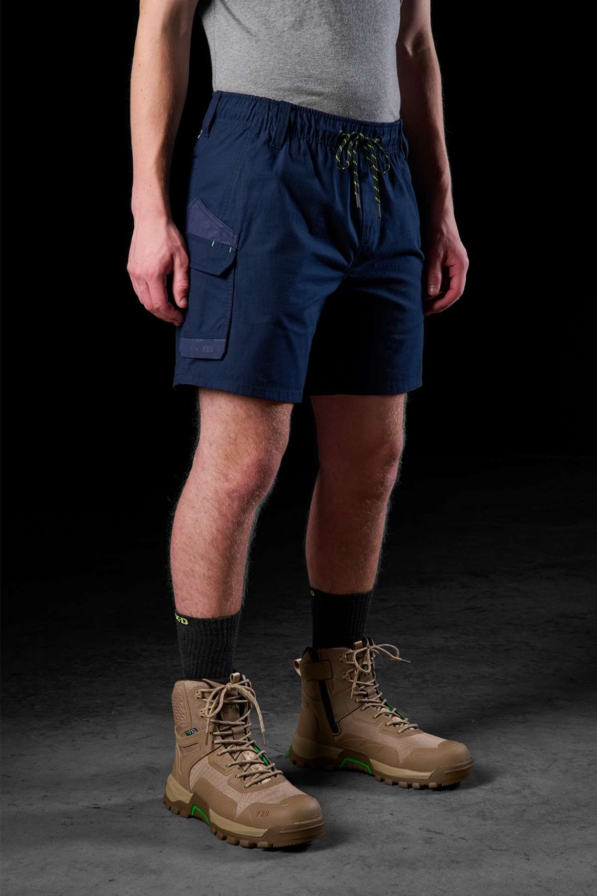 A person wearing a gray T-shirt, FXD WS-7 Work Short in durable navy blue with drawstrings and a side pocket, dark socks, and tan hiking boots stands against a dark background.