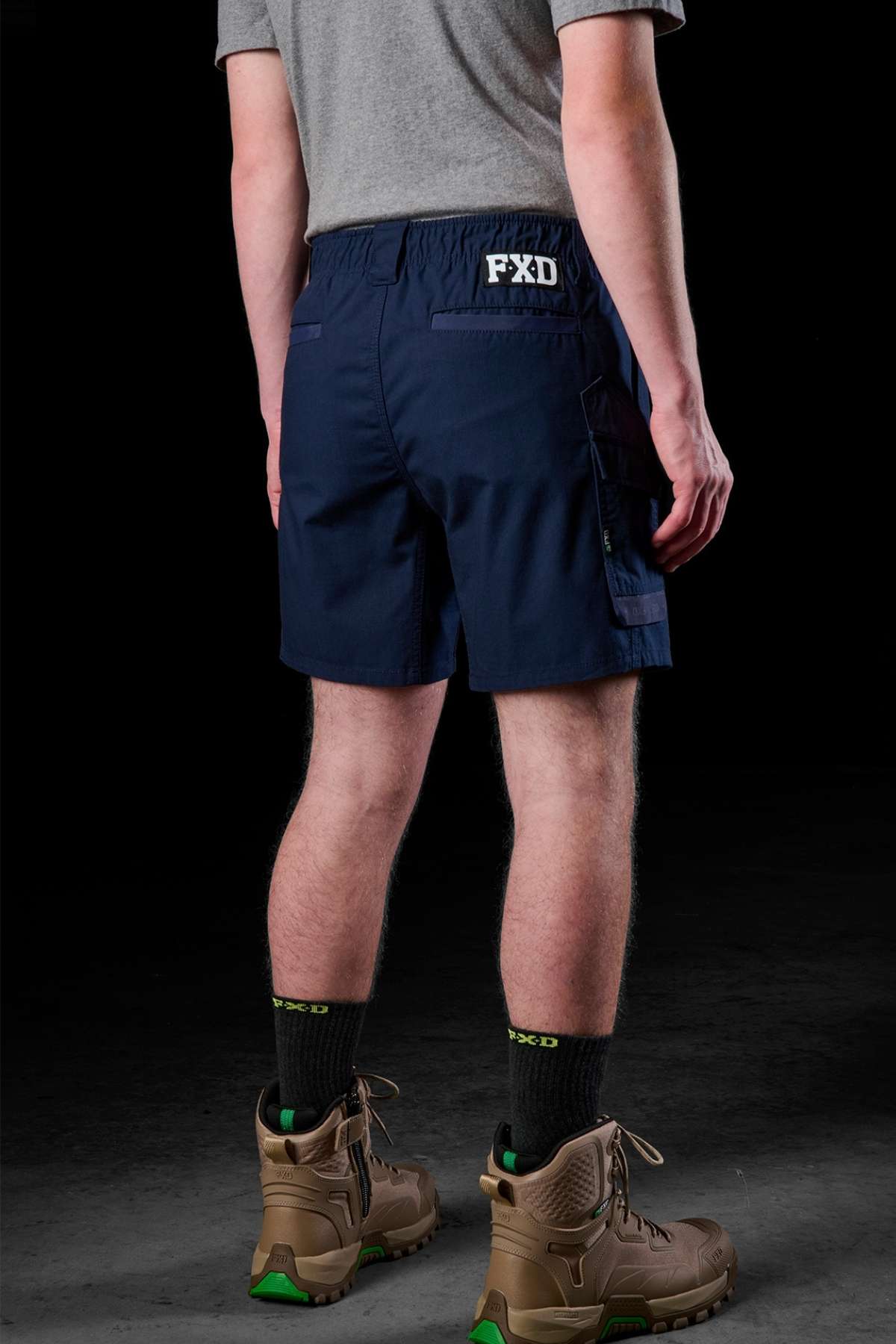 Clad in a comfortable gray t-shirt and the sturdy FXD WS-7 Work Shorts in navy blue, branded with "FXD" on the back, they complete their look with brown boots featuring green accents. Positioned against a dark backdrop, their back is turned to the viewer.