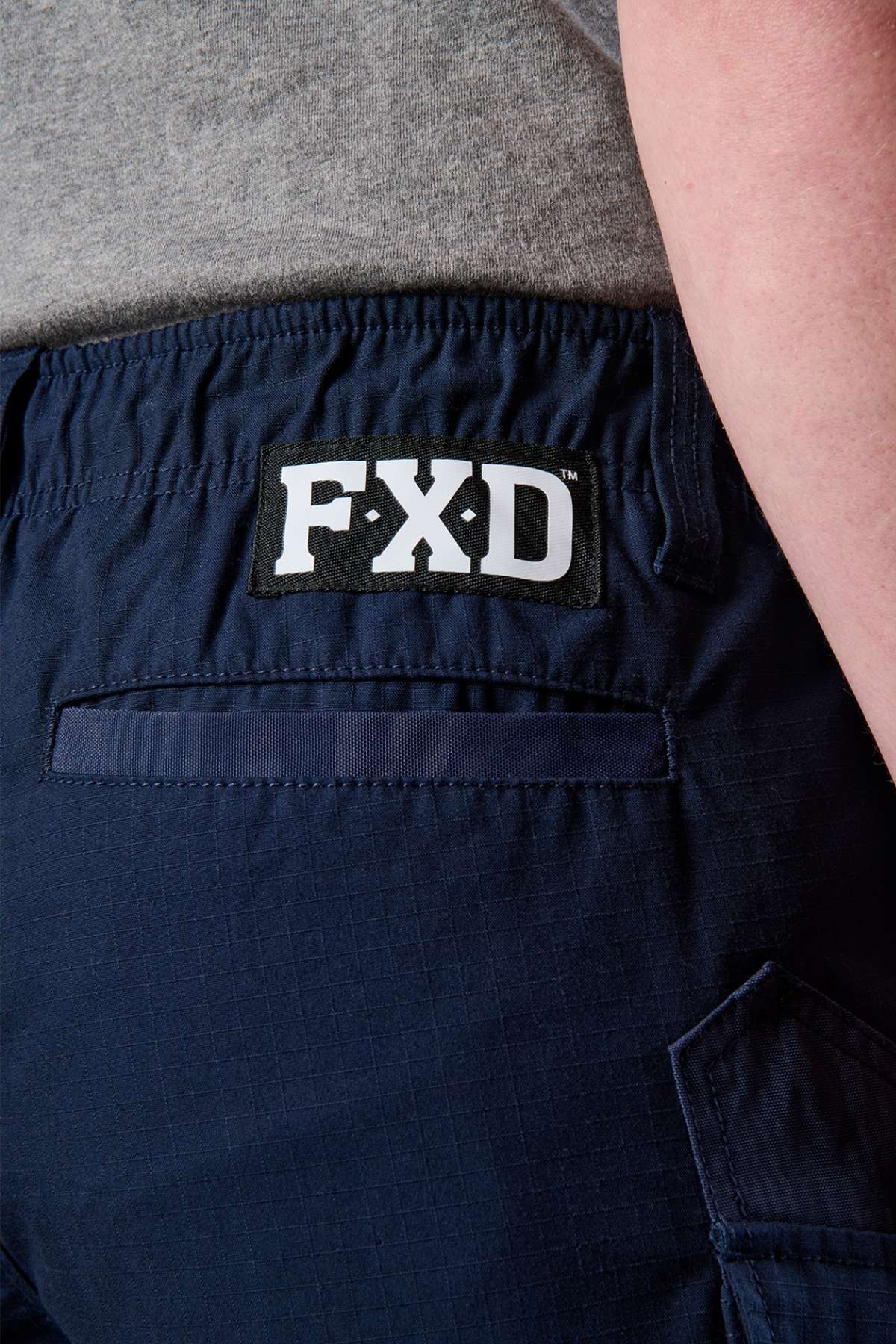 Close-up image of a person wearing the FXD WS-7 Work Short in navy blue, showcasing the "FXD" brand logo on the waistband. The shorts feature a back pocket with visible stitching, designed for comfort. The person is wearing a gray shirt tucked in.