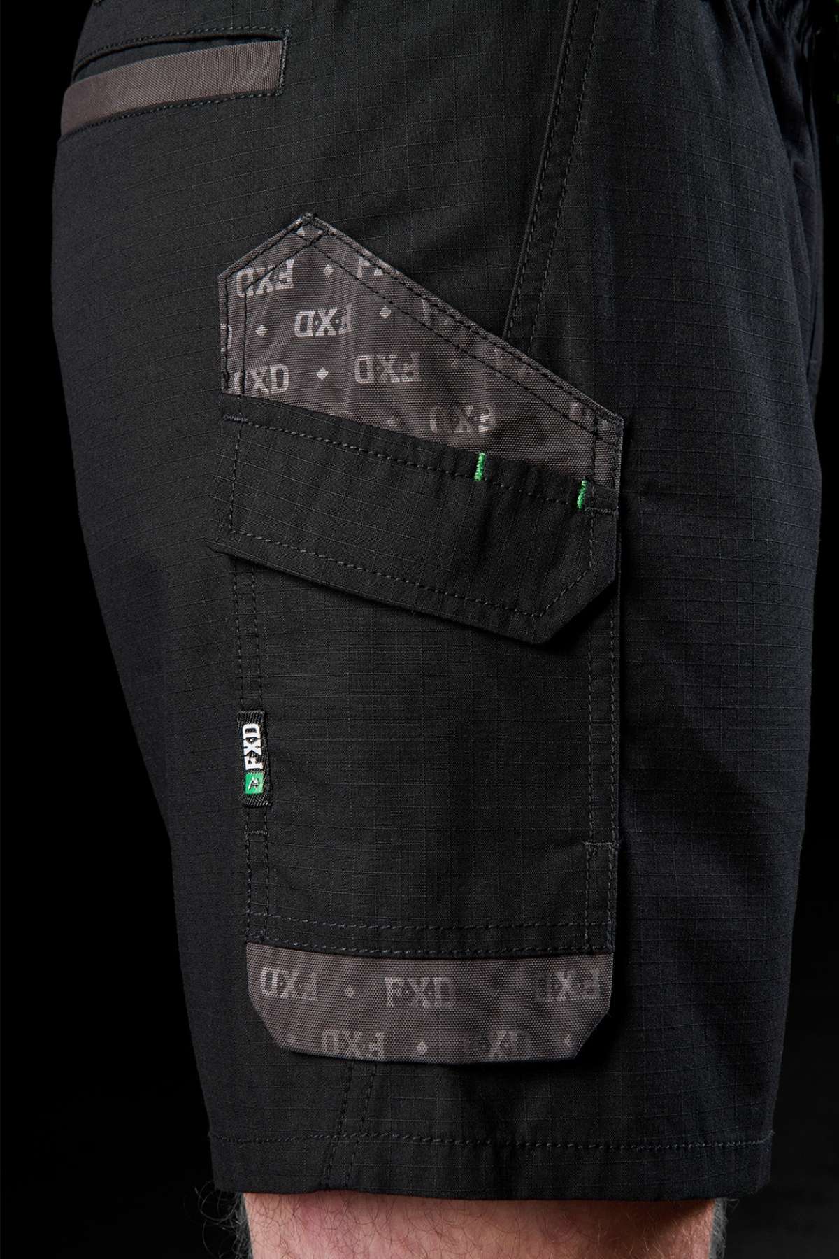 Close-up of an individual's leg sporting a black FXD WS-7 Work Short, which features an elastic waist and a pocket adorned with a patterned flap. The pocket is highlighted by small decorative green stitches, along with the "FXD" branding text around the edges. The robust fabric displays a subtle texture against the black background.